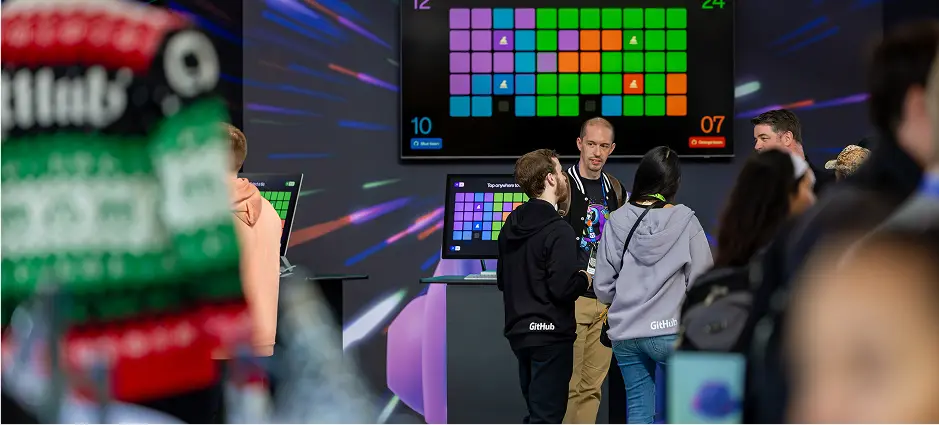 People wearing GitHub-branded clothing gather and talk near interactive screens at the Beyond Sessions area of GitHub Universe 2024. Bright, colorful grid patterns are displayed on monitors behind them, and the environment has a lively, tech-focused atmosphere with motion-themed lighting and digital visuals.